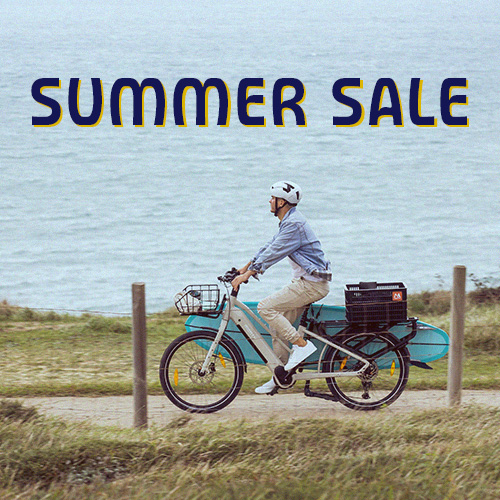 Summer Sale Monty Bikes: persona in sella a una bici cargo elettrica lungo il mare con una tavola da paddle.